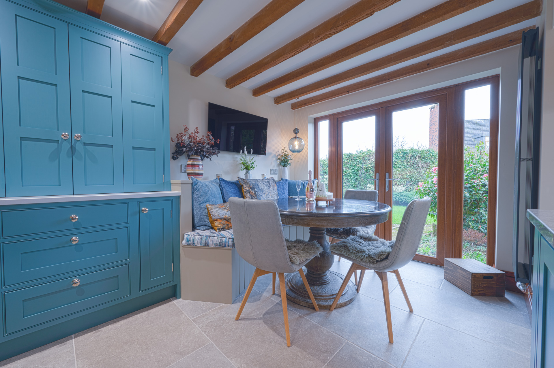 Bespoke blue handmade kitchen with built-in dining seating, exposed oak beams and garden views by Holme Tree Kitchens